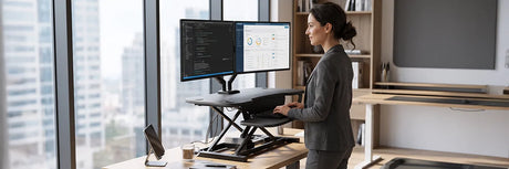 Professional woman using a black adjustable standing desk riser on a wooden desk with dual monitors, showcasing space-saving ergonomic workspace solutions.