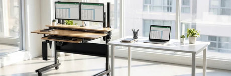 VersaDesk Foundry Bench bamboo standing desk with dual monitors next to white regular desk with laptop in bright modern office with city view