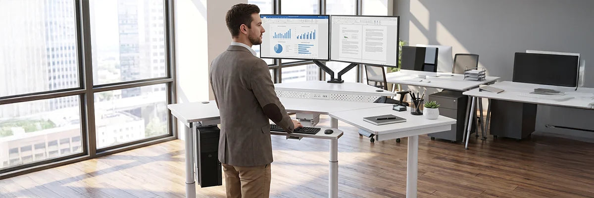 Business professional standing at an L-shaped ergonomic standing desk with dual monitors in a spacious modern office with city views.