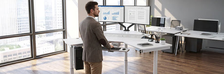 Business professional standing at an L-shaped ergonomic standing desk with dual monitors in a spacious modern office with city views.