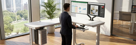 Professional using VersaDesk standing desk with organized cable management and dual monitors in modern office with city view