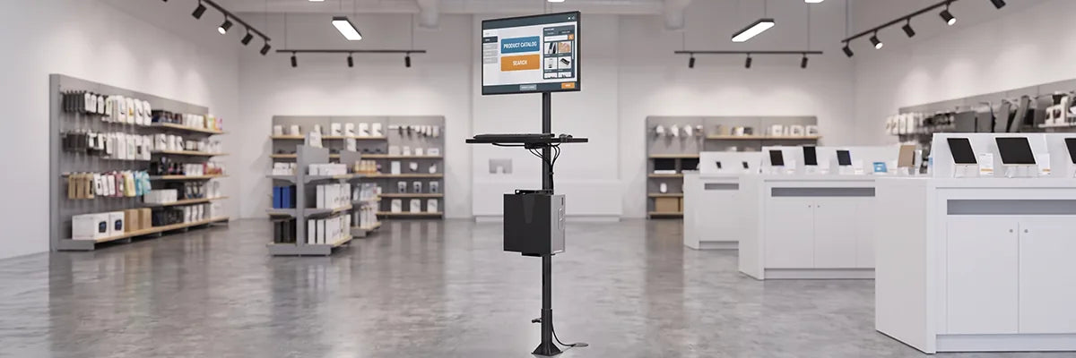 Freestanding computer kiosk stand with a touchscreen product catalog display and keyboard shelf in a bright retail store with shelving and checkout counters