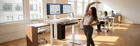 Woman stretching at her standing desk in a modern office, demonstrating easy desk exercises to stay active and reduce sedentary work habits.