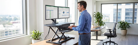 Man using a black electric standing desk riser with dual monitors on a wooden desk, comparing ergonomic desk riser options for healthier office work.