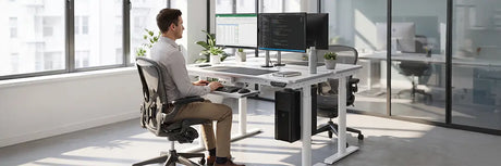 Man seated in an ergonomic chair at a height-adjustable desk with dual monitors and keyboard tray in a bright open office