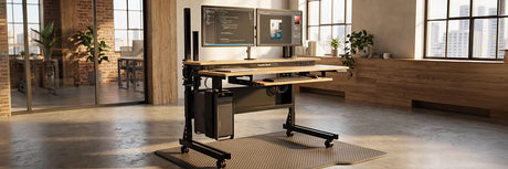 VersaDesk Foundry Bench standing desk with dual monitors on pole mount, CPU holder and anti-fatigue mat in industrial brick loft office with large windows
