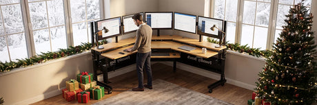 Man using a VersaDesk Foundry Bench corner standing desk with multiple monitors in a festively decorated home office with Christmas tree and wrapped gifts, perfect holiday workspace setup.