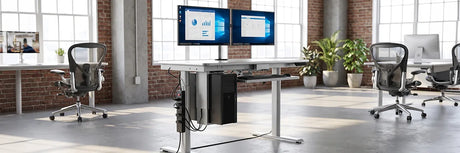 Magnetic power strip mounted under a height-adjustable standing desk with dual monitors, keyboard tray, and ergonomic mesh chairs in a bright modern office loft