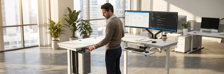 Man adjusting a white electric standing desk with dual monitors in a spacious modern office, demonstrating key considerations before buying a height-adjustable desk for work.