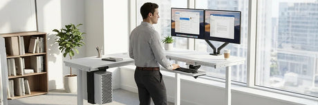Man standing at a white ergonomic standing desk with dual monitors in a clean modern office with bookshelf, showcasing the health and productivity benefits of standing workstations.