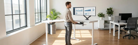 Man working at a white electric standing desk with dual monitors in a sunlit office, demonstrating how motorized adjustable desks improve physical health and reduce sedentary damage.