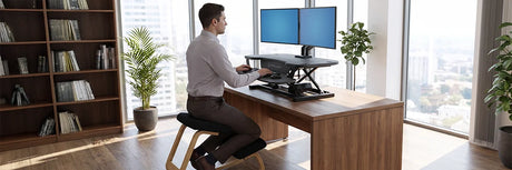 Man sitting on an ergonomic wooden kneeling chair at a desk with a black electric standing desk converter and dual monitors, demonstrating proper kneeling posture for spinal alignment in a modern office.