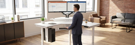 Man standing at a white L-shaped ergonomic standing desk with dual monitors in a modern office with exposed brick, demonstrating proper posture to help relieve back pain at work.