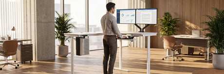 Professional man standing at white electric standing desk with dual monitors on arm mount in bright modern office with wood floors and large windows