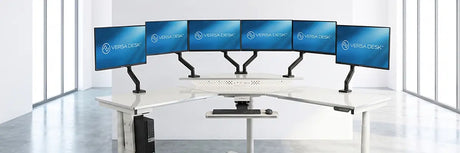 Curved white standing desk with six monitors mounted on articulating arms in a multi-monitor setup, plus a keyboard tray and CPU stand, in a bright office.