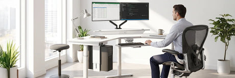 Professional seated in ergonomic mesh office chair at white VersaDesk L-shaped standing desk with dual monitors and active stool nearby