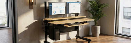 Foundry Bench standing desk with dual monitors mounted on a pole mount stand, keyboard tray, under-desk CPU holder, and potted plant in a bright room with large windows