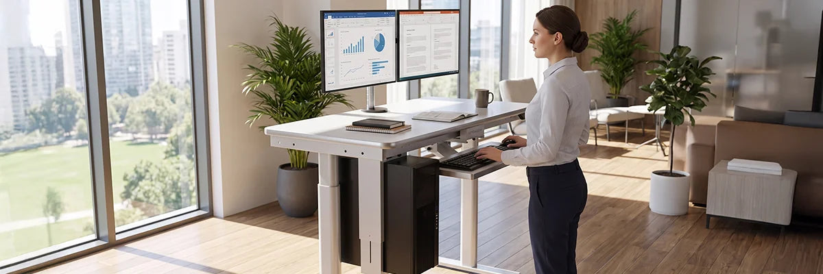 Woman working at an adjustable white standing desk with dual monitors near large windows with city skyline view, promoting office wellness and ergonomic work health.