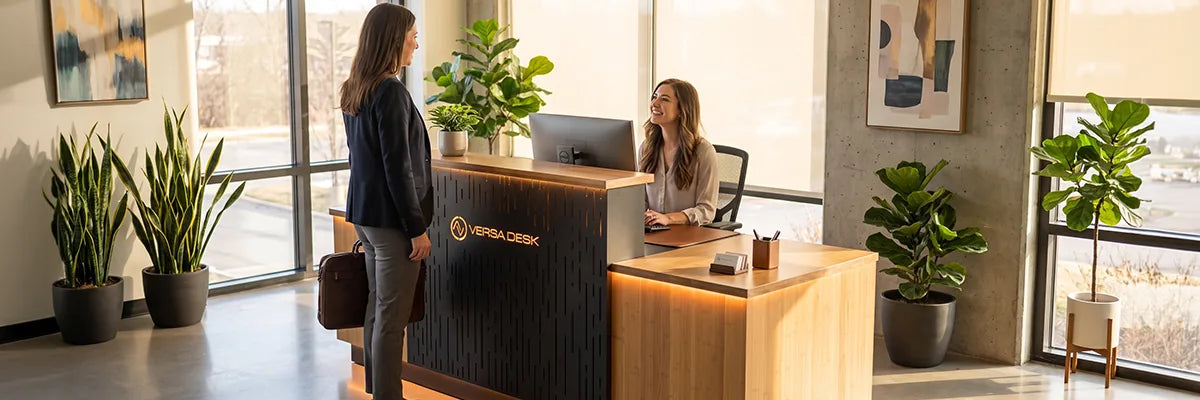 Receptionist greeting a visitor at a VersaDesk reception desk with a wood counter, black slatted front panel, potted plants, and floor-to-ceiling windows in a bright modern office lobby