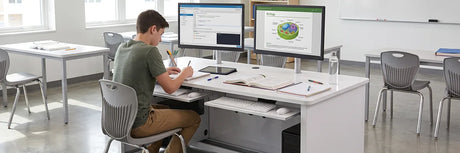 High school student working at a classroom computer desk with dual monitors displaying a biology diagram, keyboard tray, notebooks, and student chairs in a bright classroom with whiteboard
