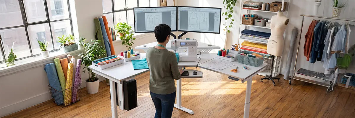 Woman standing at an L-shaped VersaDesk sewing on a machine with dual monitors showing pattern designs, fabric rolls in a sewing studio