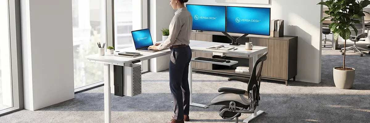 Woman standing at an L-shaped VersaDesk with laptop on the main run and dual monitors on the corner return, keyboard tray visible