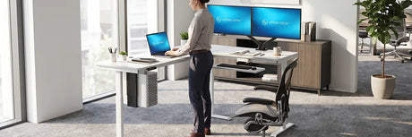 Woman standing at an L-shaped VersaDesk with laptop on the main run and dual monitors on the corner return, keyboard tray visible