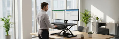 Man using a VersaDesk PowerPro standing desk converter with dual monitors on a wood desk in a conference room