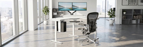 White VersaDesk standing desk with dual landscape monitors and ergonomic mesh chair in bright modern office with bookshelves and large windows