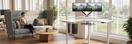 Woman working on laptop while seated in high-back grey office sofa next to white VersaDesk L-shaped standing desk with dual monitors in bright modern office