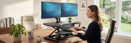 Woman working at a wooden home office desk with a black VersaDesk electric standing desk converter and dual monitors, showcasing a productive remote workspace setup with natural light.