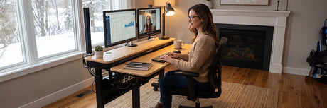 Woman working at a wooden standing desk with dual monitors in a cozy home office with a fireplace and snowy winter view through the window, showing how to stay healthy during cold months.