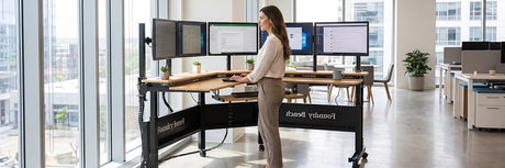 Woman standing at a large L-shaped Foundry Bench standing desk with five monitors in a bright corporate office with city views, demonstrating the health benefits of an advanced ergonomic workstation.