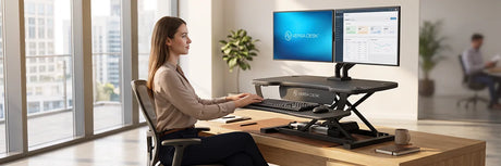 Woman sitting at a wooden desk using a black VersaDesk Power Pro electric standing desk converter with dual monitors, demonstrating the sitting position of an adjustable desktop riser in a modern office.