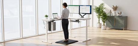 Professional woman standing on black anti-fatigue mat at white electric standing desk with dual monitors and laptop in bright modern office with floor-to-ceiling windows
