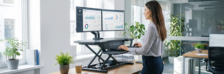 Woman standing at a black VersaDesk electric desk converter with dual monitors displaying analytics in a bright modern office, showing how small changes like standing more can improve health.