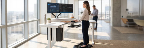 Woman standing at a white ergonomic desk with dual monitors on an anti-fatigue mat in a bright modern office with city views, demonstrating a healthy workspace setup for improved wellbeing.