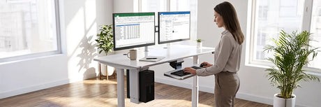 Woman working at a white ergonomic standing desk with dual monitors, under-desk CPU holder, and keyboard tray, showcasing integrated under-desk drawer and storage accessories in a bright modern office.