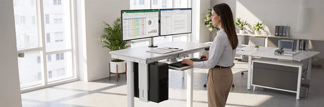 Professional woman working at a white electric standing desk with dual monitors in a bright modern office, demonstrating ergonomic workspace setup and healthy posture.