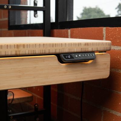Close-up of a bamboo desk edge with a digital height-adjustment control panel and warm LED accent lighting near a brick wall.