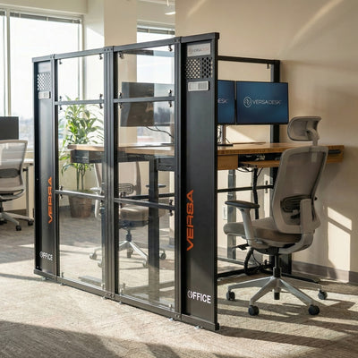 VersaDesk Versa Office WorkNest enclosed 2 person workstation pod with glass privacy panels, height-adjustable desk, dual monitors, and ergonomic chair in a bright office